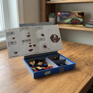 May include: A blue and gray plastic sorting tray for building blocks, featuring three internal compartments filled with assorted small plastic bricks. An open instruction manual is propped upright against the back of the tray, displaying numbered assembly steps and diagrams. The front of the tray is labeled with the text ORO.