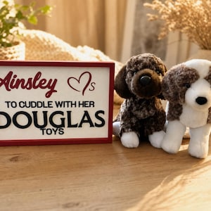 Peut inclure: Un panneau rectangulaire avec un cadre rouge portant le texte Ainsley is to cuddle with her Douglas Toys en lettres noires et bordeaux. À côté du panneau se trouvent deux chiens en peluche, l'un au pelage brun foncé et l'autre brun et blanc.
