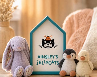 May include: A house-shaped sign with a teal frame features a black cat illustration with an orange hat and the text AINSLEY'S JELLYCATS. A lavender plush bunny with floral inner ears sits to the left, and a small penguin and duck plush sit to the right.