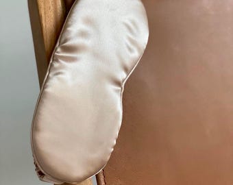 Op de afbeelding: Een lichtbeige satijnen oogmasker rust op de arm van een houten stoel met een bruine leren rugleuning. Het oogmasker is glad en lijkt gemaakt van een zacht, luxueus materiaal. De stoel heeft een modern design.
