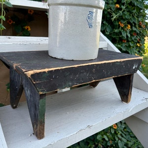 May include: A white ceramic crock with blue lettering sits on a small black and green wooden stool. The stool is on a white wooden shelf.
