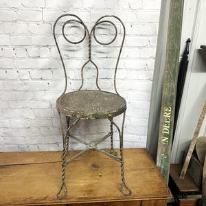 May include: An antique metal chair with a round seat and decorative back. The chair has a twisted metal design on the legs and back, and the seat appears to be made of woven material. The chair is placed on a wooden surface.