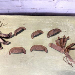 May include: A collection of 10 vintage rusty metal drawer pulls. The pulls are in various shapes and sizes, and some are bent or broken. They are all a dark brown color with rust spots.