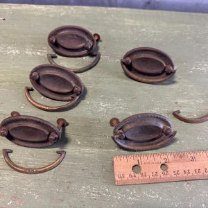 May include: Six antique oval-shaped cabinet door pulls with a brass finish and a decorative ring. The pulls are arranged on a green wooden surface with a ruler for scale.