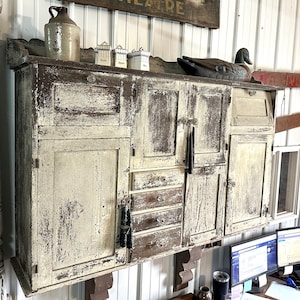 May include: A vintage white painted wooden cabinet with two doors and a drawer. The cabinet has a distressed look with visible wood grain and a duck decoy on top.