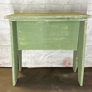 May include: A green wooden bench with a small storage compartment. The bench has a simple design with a flat top and two legs on each side.