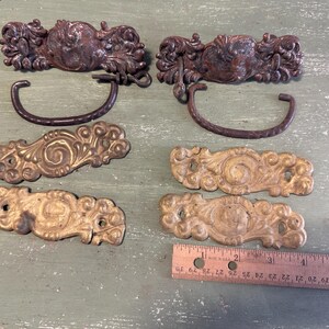 May include: Four ornate metal drawer pulls with intricate floral designs. Two pulls are made of a dark brown metal with a loop handle, and two are made of a gold metal with a flat design.