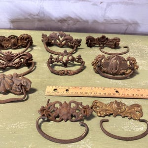 May include: A collection of antique bronze drawer pulls and hardware. The pulls feature ornate designs with curved handles and decorative backplates. A wooden ruler is included for scale. The hardware is likely for furniture restoration or vintage projects.