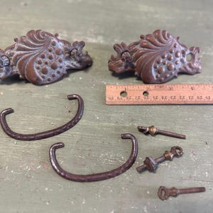 May include: Two ornate bronze drawer pulls with a decorative design, along with two matching handles, screws, and a key-shaped piece. A wooden ruler shows measurements in inches.
