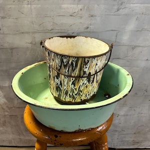 May include: A vintage green enamel basin with a brown and white marbled enamel bucket inside. The basin and bucket are sitting on a wooden stool.