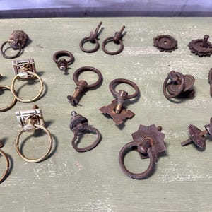 May include: Assortment of antique cabinet hardware, including drawer pulls, knobs, and hinges. The hardware is made of metal and has a variety of finishes, including brass, bronze, and nickel.