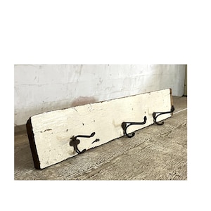 May include: A rustic, distressed white wooden coat rack with three black metal hooks. The wood has visible grain and a weathered appearance, suggesting a vintage or farmhouse style. The hooks are curved and mounted on the wooden plank.