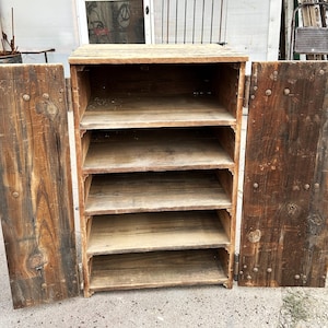 May include: A rustic wooden cabinet with two doors open, revealing five shelves. The cabinet is made of reclaimed wood with visible knots and nail heads. The wood has a warm, natural brown tone, and the cabinet appears to be handmade.