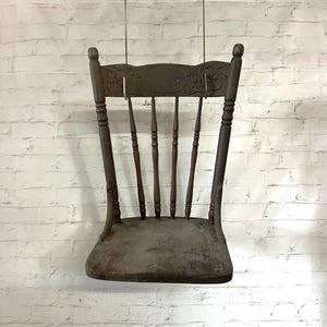 May include: An antique wooden chair with a dark brown finish. The chair features a carved backrest with floral details and a rounded seat. The chair is suspended against a white brick wall.