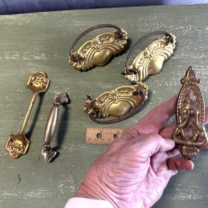May include: Assortment of antique brass drawer pulls with ornate designs.  The pulls are in various shapes and sizes, including a pair of ornate pulls with a floral design, a single pull with a more geometric design, and a pair of smaller pulls with a more simple design.