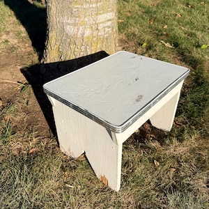 May include: A small, white wooden step stool with a light gray top and metal trim. The stool is outdoors, resting on grass near a tree. The stool is approximately 12 inches long, 10 inches wide, and 8 inches tall.