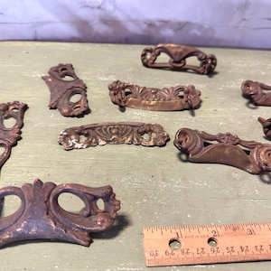 May include: A collection of eight antique ornate metal drawer pulls with intricate designs. The pulls are made of a dark, aged metal with a worn finish.