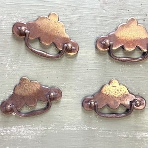 May include: Four antique brass drawer pulls with a cloud-like design. Each pull has a curved handle and is mounted with small round knobs. The pulls are set against a weathered, light green wooden surface.