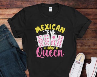 May include: Black t-shirt with the text "Mexican Train Queen" in yellow and pink, featuring dominoes and a crown graphic. The shirt is paired with blue jeans and white sneakers.