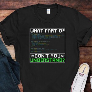 May include: Black t-shirt with the text "WHAT PART OF DON'T YOU UNDERSTAND?" in white and green. The design includes a block of code. A pair of blue jeans and white sneakers are visible.