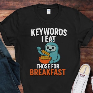 May include: Black t-shirt with the text "KEYWORDS I EAT THOSE FOR BREAKFAST". The shirt features a cartoon robot character holding a bowl with "keyword" labels. A pair of blue jeans and white sneakers are visible.