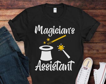 May include: Black t-shirt with white text that reads "Magician's Assistant". The design includes a white top hat, a yellow magic wand, and yellow stars.