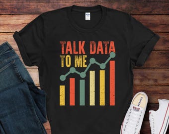 May include: Black t-shirt with the text "TALK DATA TO ME" in distressed orange and yellow lettering. Below the text is a graphic of a line graph and bar chart in green, orange, yellow, and teal. The shirt is paired with blue jeans and white sneakers.