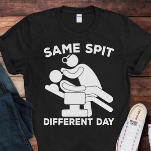 May include: Black t-shirt with the text "SAME SPIT DIFFERENT DAY" and a white graphic of a dentist and patient. The shirt is displayed with blue jeans and white sneakers on a wooden surface.