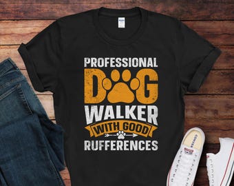 Op de afbeelding: Zwart T-shirt met de tekst "PROFESSIONAL DOG WALKER WITH GOOD RUFFERENCES". Het woord "DOG" staat in grote oranje letters met een pootafdrukontwerp. Het shirt wordt gecombineerd met een blauwe jeans en witte sneakers.