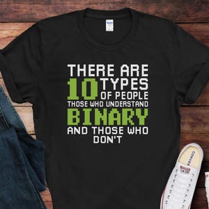 May include: Black t-shirt with white and green text that reads "THERE ARE 10 TYPES OF PEOPLE THOSE WHO UNDERSTAND BINARY AND THOSE WHO DON'T." The shirt is paired with blue jeans and white sneakers.