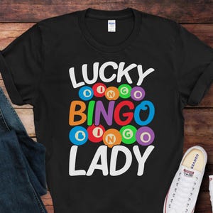 May include: Black t-shirt with the words "LUCKY BINGO LADY" in white, with colorful bingo ball graphics. The shirt is paired with blue jeans and white sneakers.