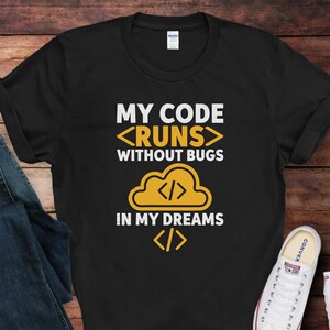 May include: Black t-shirt with the text "MY CODE <RUNS> WITHOUT BUGS IN MY DREAMS" in white and yellow. The design includes a cloud graphic with code symbols. A pair of blue jeans and white sneakers are visible.