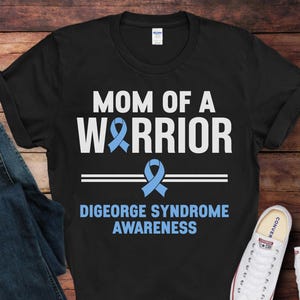 May include: Black t-shirt with white text that reads "Mom of a Warrior" and a blue ribbon. Below the text is "DiGeorge Syndrome Awareness".
