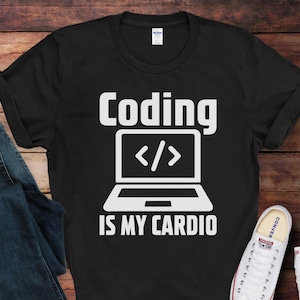 May include: Black t-shirt with the text "Coding IS MY CARDIO" in white. A white laptop graphic with code symbols is centered below the word "Coding". Blue jeans and white sneakers are visible in the image.