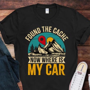 May include: Black t-shirt with a graphic design featuring mountains, a red location pin, and the text "FOUND THE CACHE, NOW WHERE IS MY CAR". The shirt is paired with blue jeans and white sneakers.