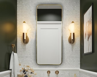 Puede incluir: Un baño moderno con un lavabo blanco, grifos dorados y un mueble de tocador de madera oscura. Las paredes están alicatadas con pequeñas baldosas blancas y el espejo tiene un marco dorado. Apliques decorativos flanquean el espejo y una toalla cuelga de un anillo dorado.