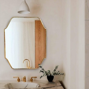 May include: A white porcelain sink with gold faucets sits on a marble countertop with a wooden cabinet underneath. A gold framed octagonal mirror hangs above the sink. A small vase with white flowers sits on the countertop.
