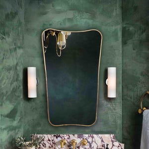 May include: A bathroom interior with a marble countertop and gold fixtures. A large mirror with a gold frame hangs above the sink, flanked by two cylindrical wall sconces. The walls are painted a textured green. The sink is made of brown metal.