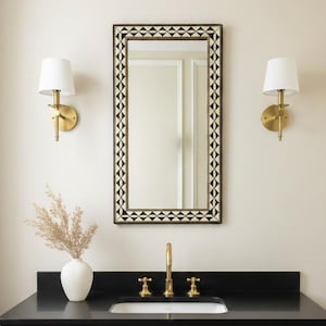 May include: A modern bathroom vanity with a black countertop and a gold faucet. A rectangular mirror with a geometric black and white border hangs above the sink. Two gold sconces with white shades flank the mirror. A white towel hangs from a gold bar.