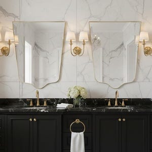 May include: A luxury bathroom vanity featuring black cabinets, a black marble countertop, and gold fixtures. Two uniquely shaped mirrors with gold frames are mounted above the vanities. The walls are white marble. A white towel hangs from a gold ring.