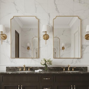 Könnte beinhalten: Ein dunkelbrauner Doppelwaschtisch mit goldenen Beschlägen und einer Marmorarbeitsplatte. Zwei goldgerahmte Spiegel hängen darüber, flankiert von Wandleuchten mit weißen Schirmen. Der Hintergrund zeigt weiße Marmorfliesen und einen Fischgrätboden.