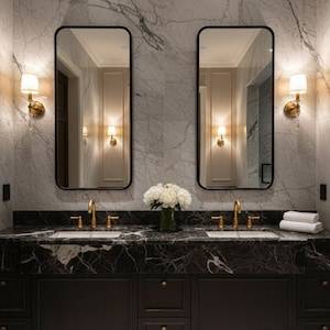 Puede incluir: Un lujoso mueble de baño con encimera de mármol negro y gabinetes de madera oscura. Dos espejos rectangulares con marcos negros cuelgan sobre los lavabos, y los accesorios dorados añaden elegancia. Flores blancas y toallas completan el diseño.