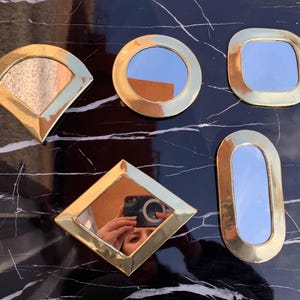 Puede incluir: Cinco espejos decorativos de color dorado con diferentes formas y tamaños. Los espejos están sobre una superficie de mármol blanco y negro.