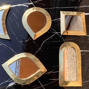 Peut inclure: Cinq miroirs décoratifs dorés de formes et de tailles différentes. Les miroirs sont posés sur une surface en marbre noir.