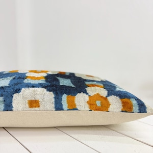 May include: Decorative pillow with a geometric pattern in shades of navy blue, light blue, white, and orange. The pillow has a beige base and a soft, textured appearance, suitable for home decor.