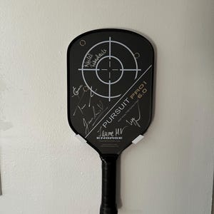 May include: Black pickleball paddle with a target design and the text "PURSUIT PRO 6.0". The paddle has a black grip and is signed by multiple people. The brand name "ENGAGE" is also visible.