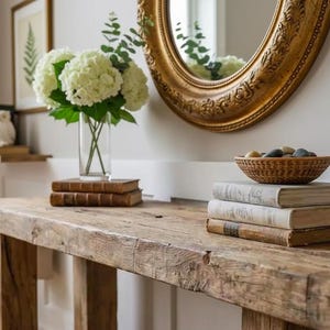 Op de afbeelding: Een rustieke houten console met een verweerde afwerking. Het heeft een heldere glazen vaas met witte bloemen, gestapelde vintage boeken, een geweven kom met stenen en een goudomrande spiegel.