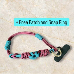 Könnte beinhalten: Ein handgefertigtes Handy-Lanyard in Burgunderrot und Türkis. Das Lanyard hat ein gewebtes Design mit türkisfarbenen Akzenten und einem blütenartigen Knoten. Es enthält einen Schnappring und einen schwarzen Handygriff.