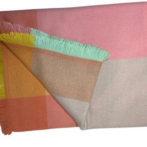 May include: A colorful woven blanket with a fringed edge. The blanket features a patchwork design with squares of pink, peach, yellow, and mint green. The blanket is folded over, revealing the different color blocks.