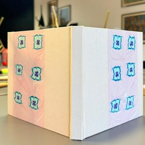 Puede incluir: Un libro cuadrado hecho a mano con una cubierta de color melocotón claro y una tira decorativa de papel veteado rosa y blanco. La tira está adornada con diseños florales turquesa y morados. El libro está sobre una superficie gris.